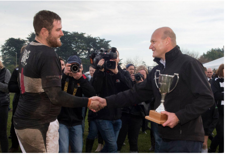Edward Luisetti, presents the Luisetti Seeds Combined Country Cup to Waihora Captain; Matthew Stone. Matthew's clothes are muddy but he is smiling widely. In the background are a crowd of rugby fans.
