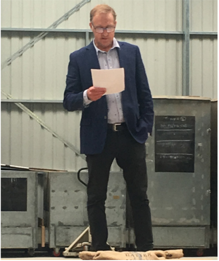 Image of Vincent Luisetti, reading from a paper in his hand while stood on a stage. The wall behind him is corrugated steel, suggesting the function took place in a barn or warehouse